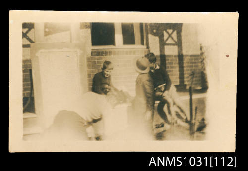 Black and white photograph of four people sitting on a porch in a group, or on the ground in front of the porch