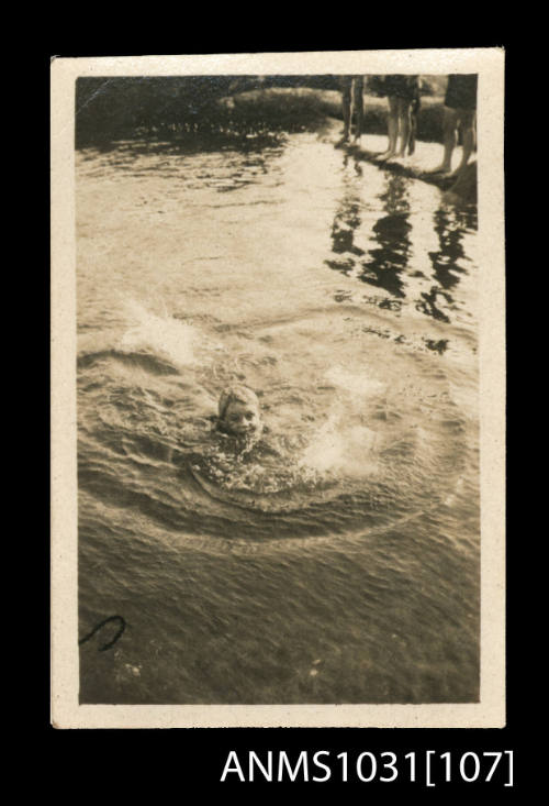 Young boy splashing in the water, with peoples legs visible in the top right corner