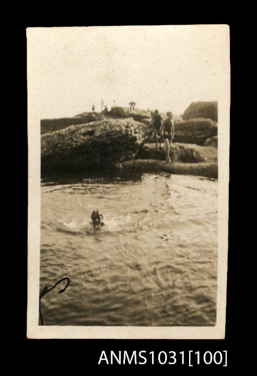 Young boy swimming in the water next to the shoreline, with several people standing on large rocks beside the water