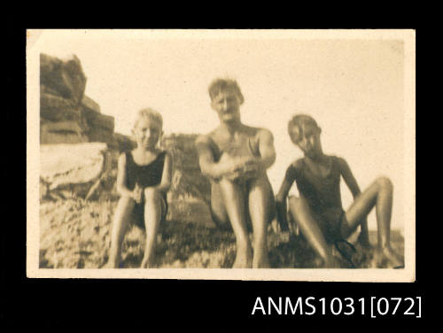 Man and two young boys sitting on a rocky section of beach in their swim suits