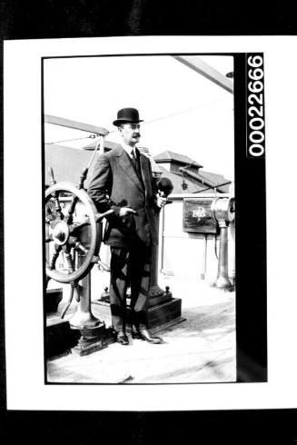 Man in suit and bowler hat, standing beside a small ship's wheel