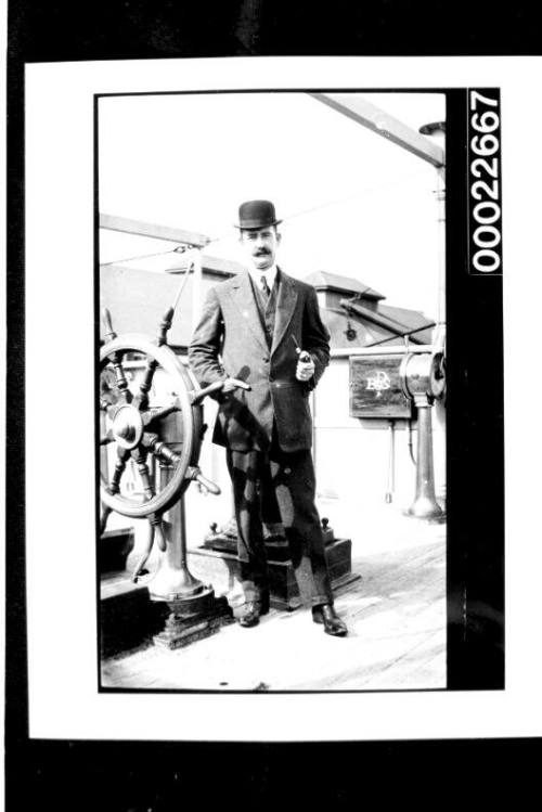 Man in suit and bowler hat, standing beside a small ship's wheel on deck