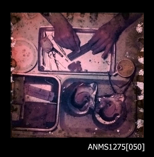 Colour negative of three containers, one of which contains two pearl shells, surrounded by pearl seeding equipment, and a mans' hand holding an instrument