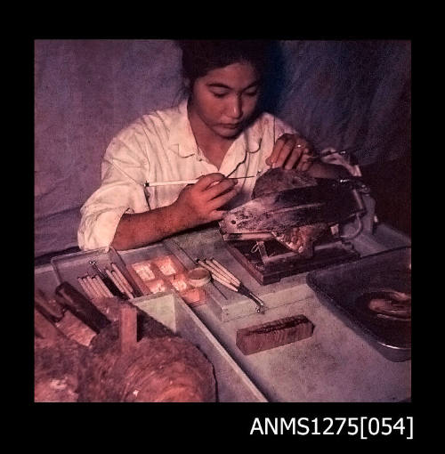 Colour negative of Yurie (or Yulie) George sitting at a desk, using dental tools to seed a pearl shell