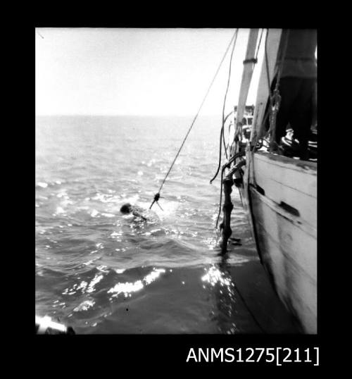 A man in the water, wearing a diving helmet, holding onto rope over the edge of a lugger