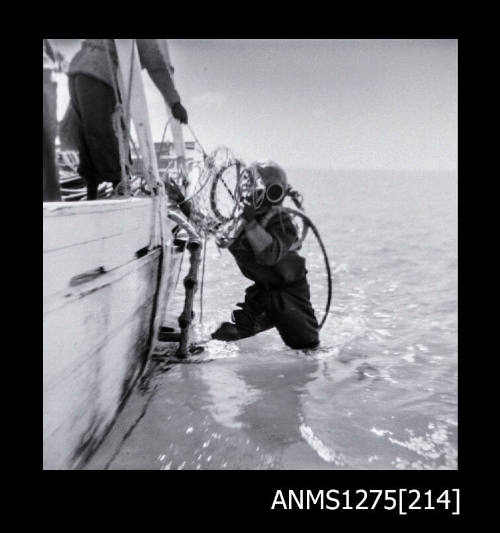 A man in a diving suit and helmet, hanging over the side of a lugger, partially in the water