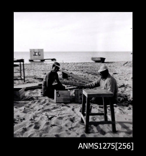 Two men sittibng on the beach, with equipment on the sand and a pearl raft and a boat in the water in the background