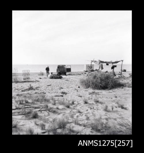 A car, shade cover, equipment and several people on a beach, with a pearl raft in the water in the background