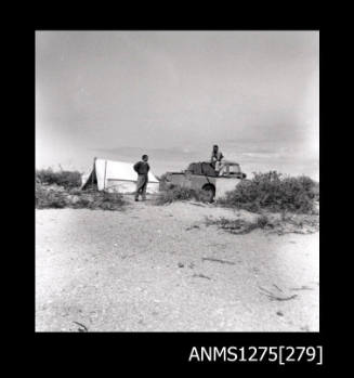 A man standing in a boat, and a man standing in front of a tent, on the sand