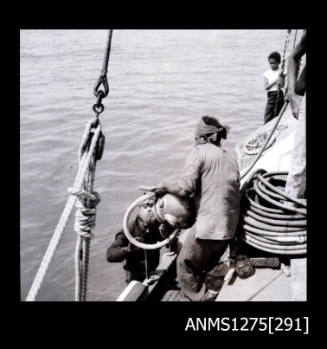 Several people on a lugger, with two men pulling a diver out of the water and over the edge of the lugger