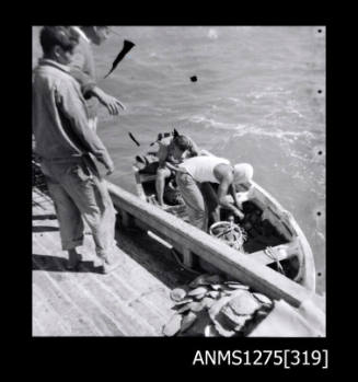 Two people standing on the side of a boat, looking into a smaller boat with two men lifting pearl shell baskets