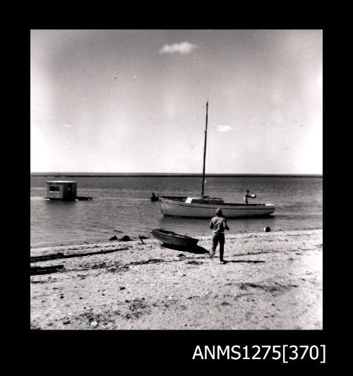 A person standing on the shoreline near a boat, and in the water in the background is a pearl raft, possibly being pulled by a boat