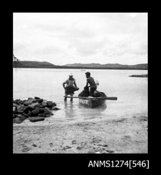 Two men and a boat, one standing in the boat, and the other standing in shallow water, on Packe Island