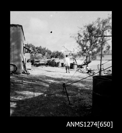 A man walking on the shoreline near palm trees, equipment and a shed, on Packe Island