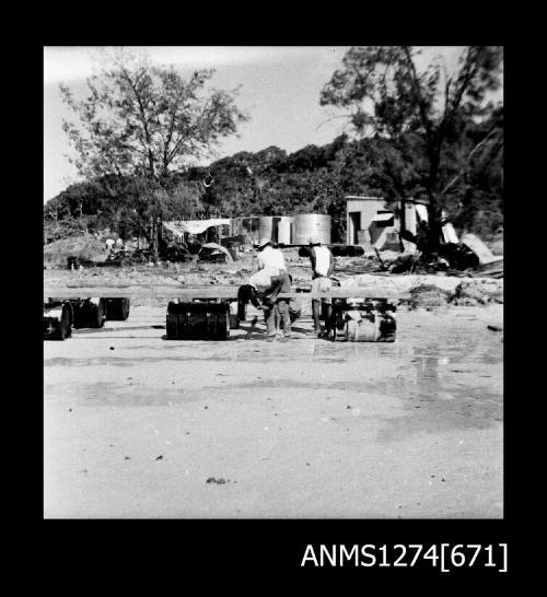 Three men, constructing the base of a lab from wood and cylindrical containers, on Packe Island