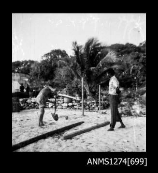 Two men on the beach, one standing next to wooden planks, the other digging a hole, on Packe Island