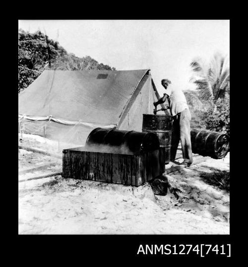 A man standing in front of a tent, with a metal drum on his left hand side in a crate, on Packe Island