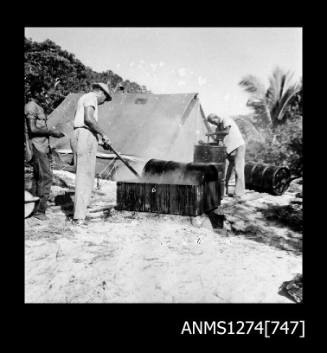 Three men working with metal drums, one of which is in a wooden crate, on Packe Island