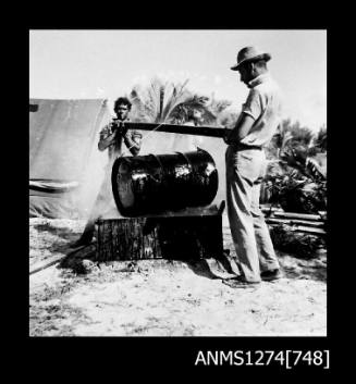 Two men holding a wooden beam over a metal drum on Packe Island