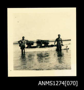 A small boat in shallow water, with two men standing in front of it, and with wire pearl cages sitting on the top of it, on Packe Island