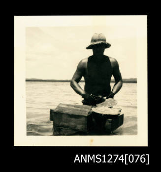 A man, standing in the water, and holding a pearl shell on a wooden block/table, on Packe Island