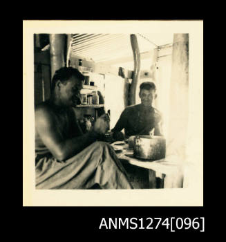 Two men (one of which is Denis George) sitting at a desk, inside of a room, or shed, on Packe Island