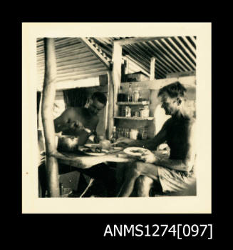 Two men (one of which is Denis George) sitting at a desk, inside of a room, or shed, on Packe Island