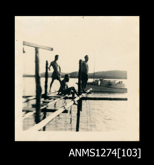 Three men sitting and standing on a wooden pearl raft, with a boat and island in the background, on Packe Island