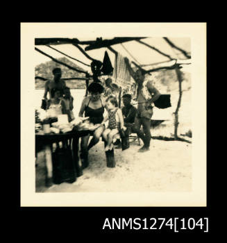 Photograph of several men, women and children, sitting underneath a shade structure on a beach, at a table, on Packe Island