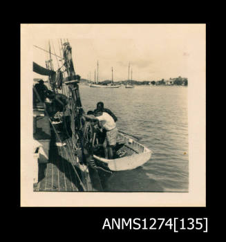 Two men in a boat, which is floating next to a larger boat, with small boats visible in the background, on Packe Island