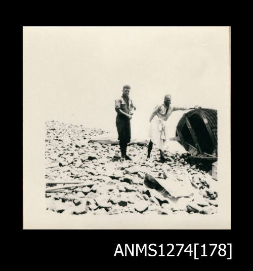 Two men standing on rocks, one of whom is holding up an overturned boat, with captured sea mammal, possible a dugong, lying on the rocks in the background, on Packe Island