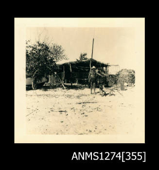 Shack on a beach, with a man (possibly Denis George) standing in front it alongside two dogs, on Packe Island