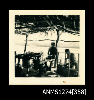 Two men (one is Denis George), sitting under a shade structure on the beach, with various pieces of equipment, on Packe Island