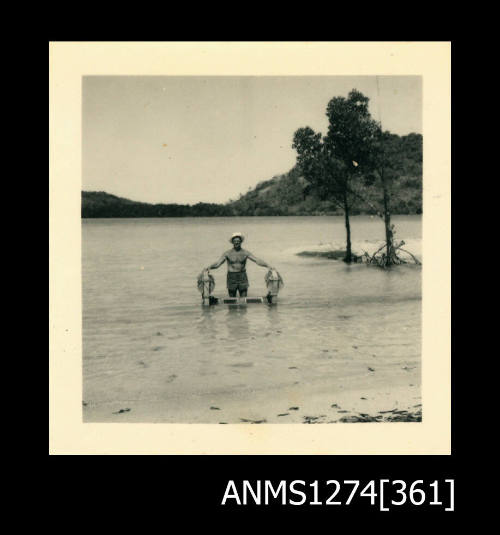 A man standing in shallow water, holding the wooden object from ANMS1274[359], on Packe Island