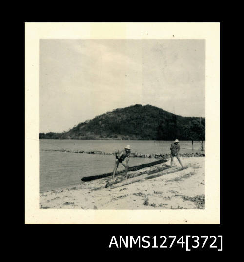 A two men standing on a beach (the man on the left is Denis George), with mangrove tree logs, on Packe Island