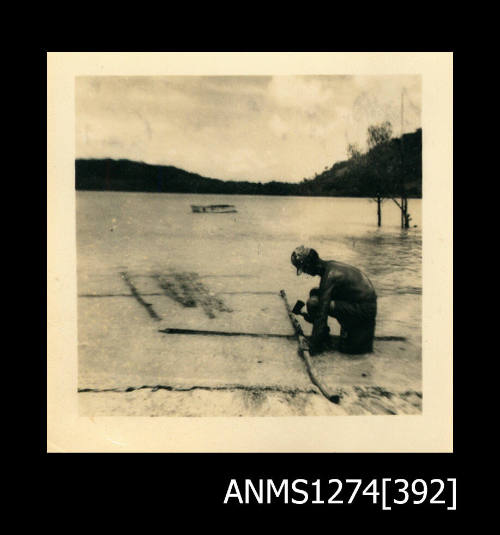 A man sitting in shallow water on a beach, holding an axe, with two crossed logs, on Packe Island