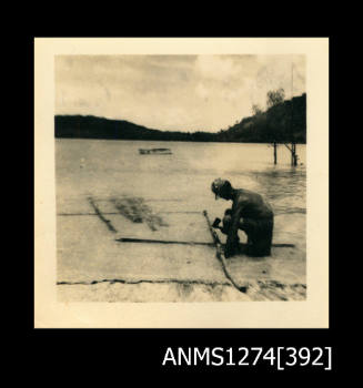 A man sitting in shallow water on a beach, holding an axe, with two crossed logs, on Packe Island