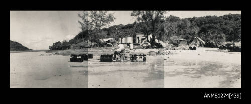 A beach with sheds, tents, trees and equipment, and three men working on metal containers with wooden beams, on Packe Island