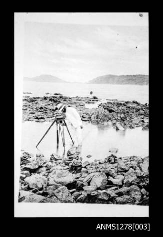 A man in a suit, standing in shallow water, with a tripod shaped object
