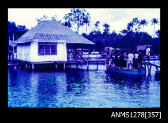 35mm colour transparency of a a shed over the water, next to which is a small boat filled with numerous standing people