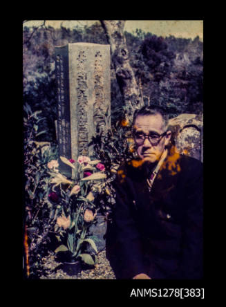 35mm colour transparency of a Japanese man standing next to Mikimoto's tombstone, decorated with flowers