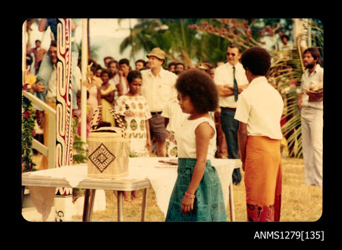 Young girl, wearing a blue skirt and white top, standing next to a table with a basket, during their Queen Elizabeth II and Prince Philip's visit to Papua New Guinea in 1977