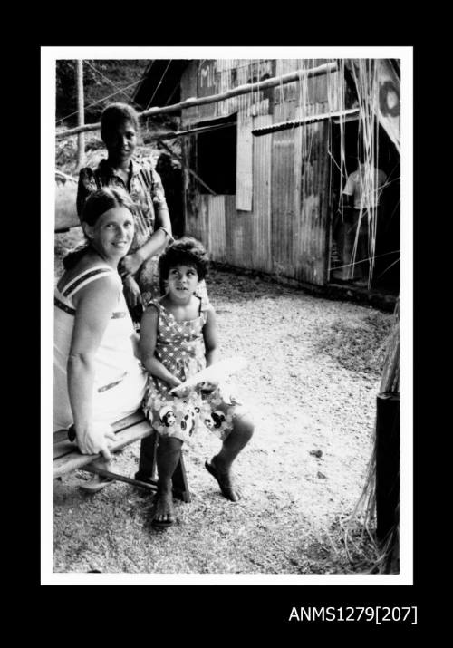 A European woman sitting on a chair, next to two Papua New Guinean girls, next to a shed