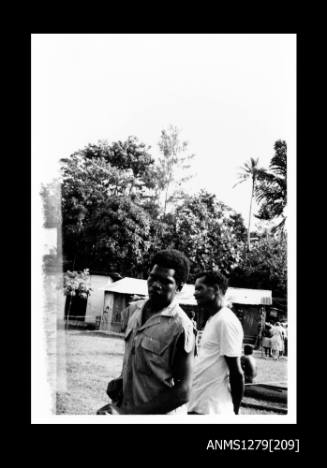 Two Papua New Guinean men standing outdoors in front of a house or shed