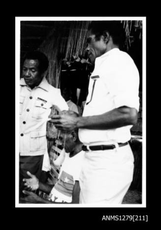 Three Papua New Guinean men, two standing and one sitting, with the man in the forefront holding a piece of pearl shell jewellery