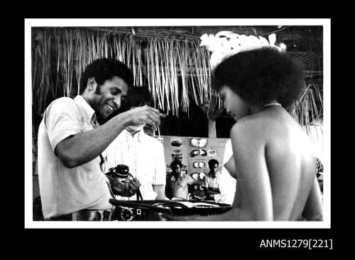 A Papua New Guinean girl in traditional dress, holding a tray containing pearl or pearl shell jewellery, and a Papua New Guinean man holding a camera, who is picking up a piece of the jewellery