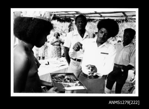 A Papua New Guinean girl in traditional dress, holding a tray containing pearl or pearl shell jewellery, and a Papua New Guinean man picking up a piece of the jewellery