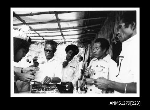 A Papua New Guinean girl in traditional dress, holding a tray containing pearl or pearl shell jewellery, and four Papua New Guinean men looking at and holding pieces of the jewellery