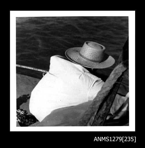 The back of a man, who is sitting on the edge of a boat, wearing a white shirt and straw hat