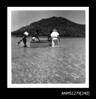 A man wearing white clothes and a straw hat, and two Papua New Guinean men, walking in shallow water beside a dinghy